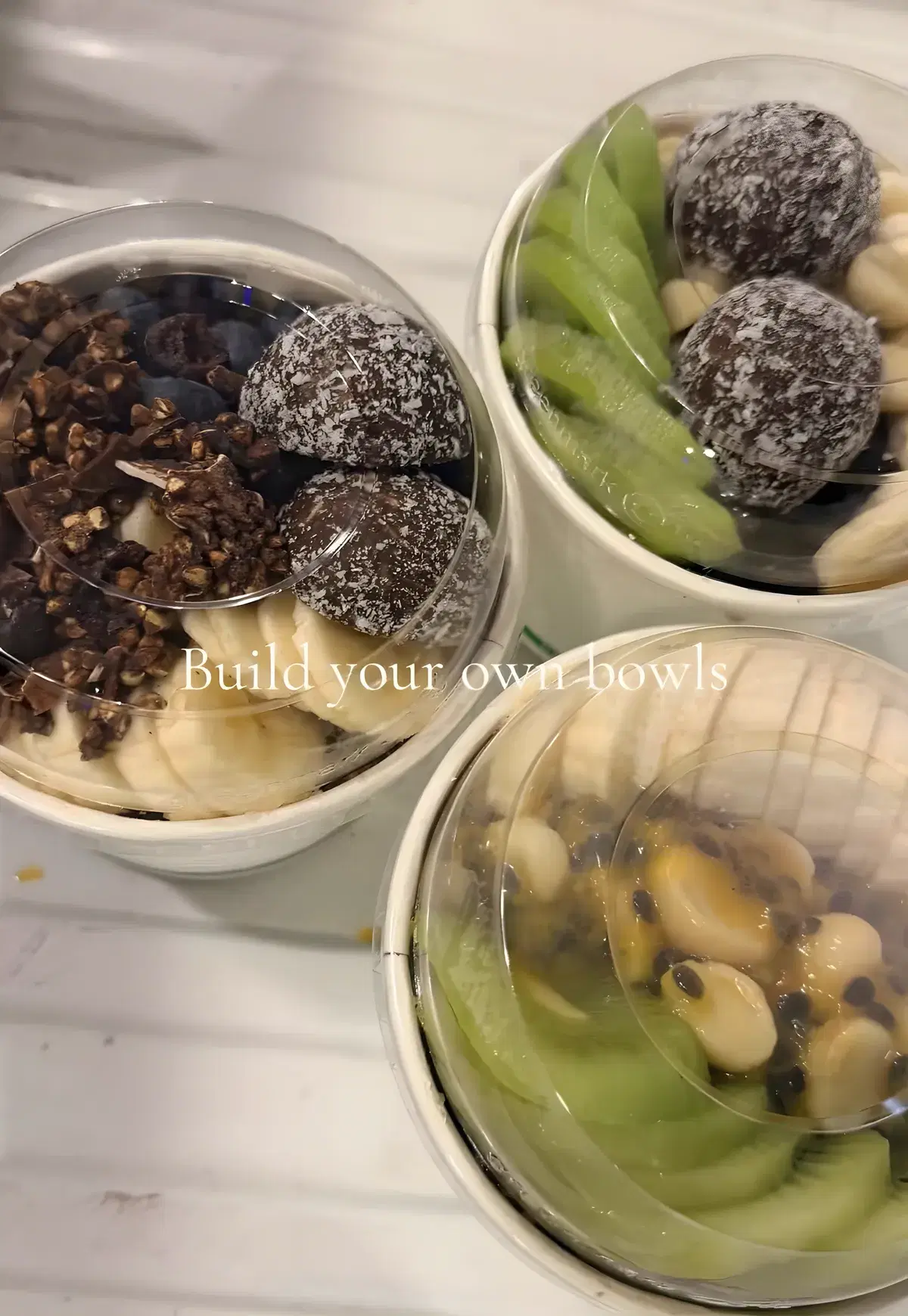 Three lidded build-your-own acai bowls topped with kiwi fruit, banana, bliss balls and granola